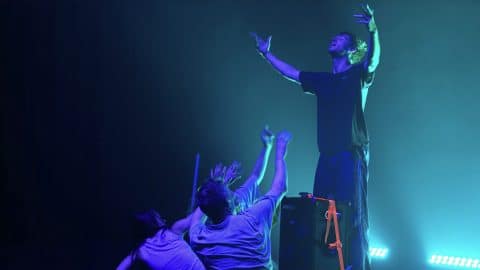 An image from BPM with dramatic blue-green lighting. One performer stands elevated with arms stretched upward and face lifted, while three others reach up toward him from below, creating a powerful, almost worship-like tableau.