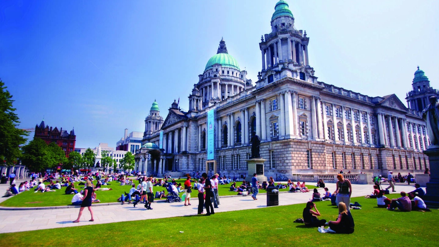 Belfast City Hall in summer, with groups of people sitting on the lawns enyoing the sunshine. Courtesy of Tourism NI