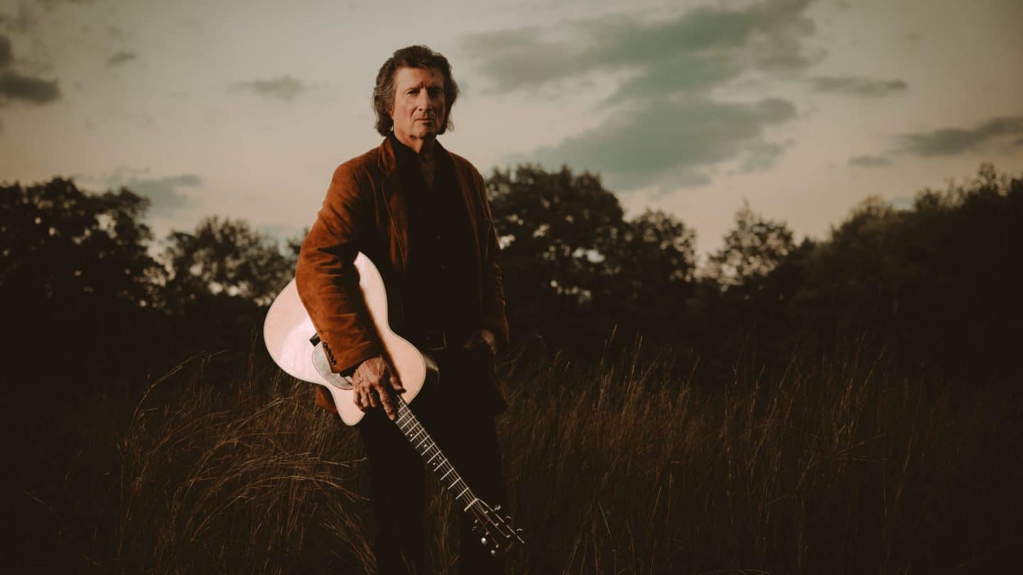 Chris Smither, holding his guitar, standing in a field, with trees in the background.