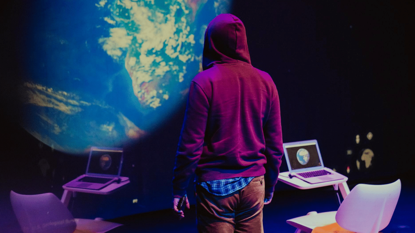 DOING IT. Photographer Ana Viotti. Image of the earth taken from space, displayed as stage backdrop. In middle foreground, a man dressed in a maroon coloured hoodie looks at the large image of the earth. in the bottom left and right corners, a laptop displays the same image. The laptop sits on a desk, along with a white chair
