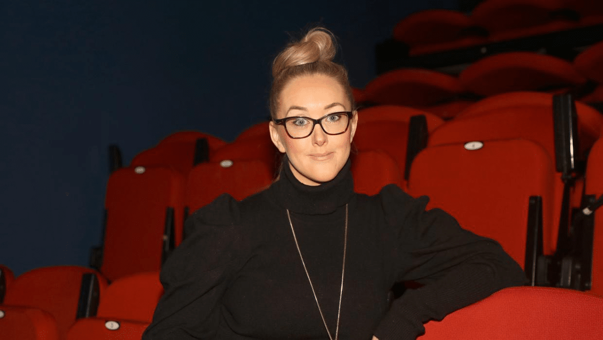 Eileen McClory, who is seated in a theatre auditorium. She is wearing a black jumper, long gold chain and black rimmed glasses.