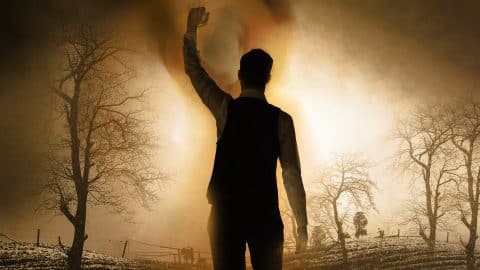 A silhouetted figure stands with one arm raised against a glowing, sepia-toned landscape of bare winter trees and misty light.