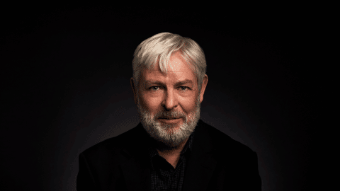Jonathan Coe is a middle aged white man, with grey hair and a short grey beard. He wears a black shirt.