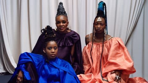 Three members of Les Amazones d'Afrique in colourful traditonal gowns, against a grey backdrop.