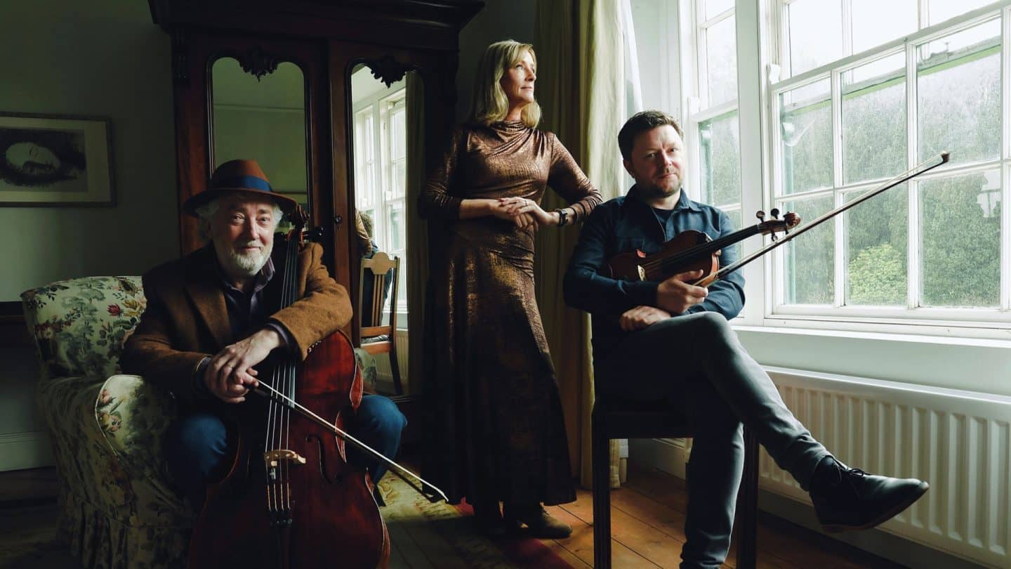Musicians Neil Martin, Mary Dillon and Dónal O’Connor are pictured in a softly lit room: Martin sits with his cello, Dillon stands by the window gazing out, and O’Connor sits nearby holding his violin.