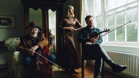 Musicians Neil Martin, Mary Dillon and Dónal O’Connor are pictured in a softly lit room: Martin sits with his cello, Dillon stands by the window gazing out, and O’Connor sits nearby holding his violin.