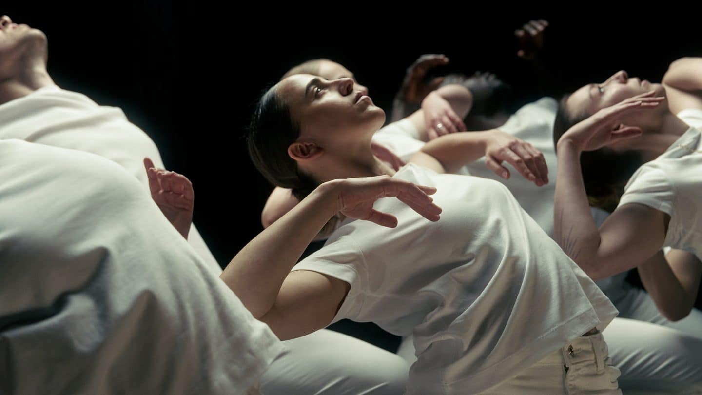A group of dancers dressed in white move in unison against a dark stage background. In the foreground, a performer arches backward with arms softly raised and gaze lifted upward.