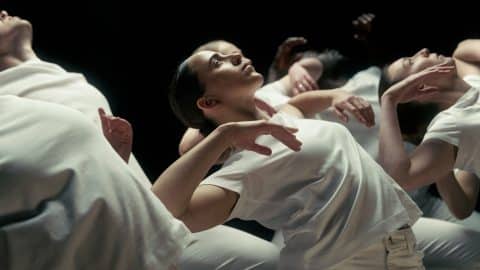 A group of dancers dressed in white move in unison against a dark stage background. In the foreground, a performer arches backward with arms softly raised and gaze lifted upward.
