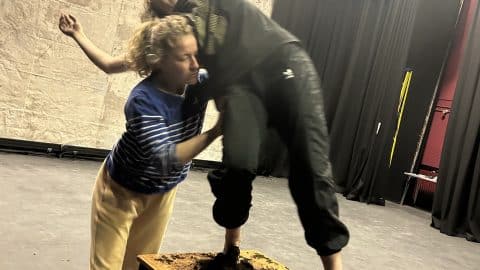 Two female dancers in rehearsals for ROOTS. One is standing on a table, with her feet hidden by brown soil. She appears to be about to fall but is supported by the second person leaning into her.