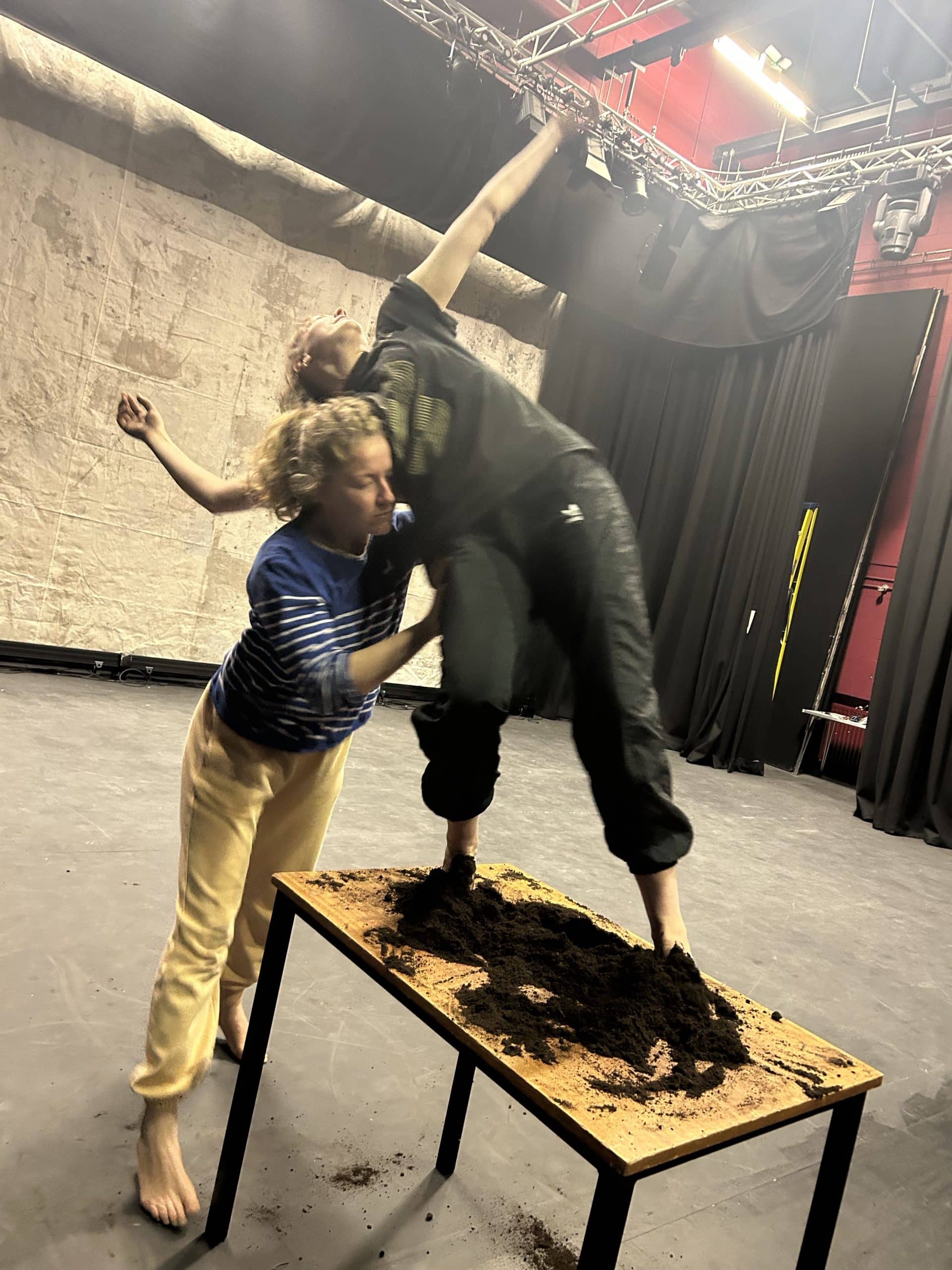 Two female dancers in rehearsals for ROOTS. One is standing on a table, with her feet hidden by brown soil. She appears to be about to fall but is supported by the second person leaning into her.