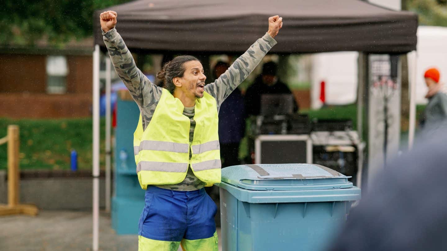 Circus artist Said Mouhssine, wearing a Hi-Viz vest, holds his arms aloft in triumph. There is a blue recylcing bin beside him.