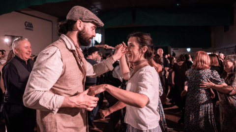 A bearded man in a flat cap and waistcoat dances with a smiling woman in a white top, holding her hand as they move together. Behind them, a crowded dance hall is filled with people dancing and chatting under warm, low lighting.