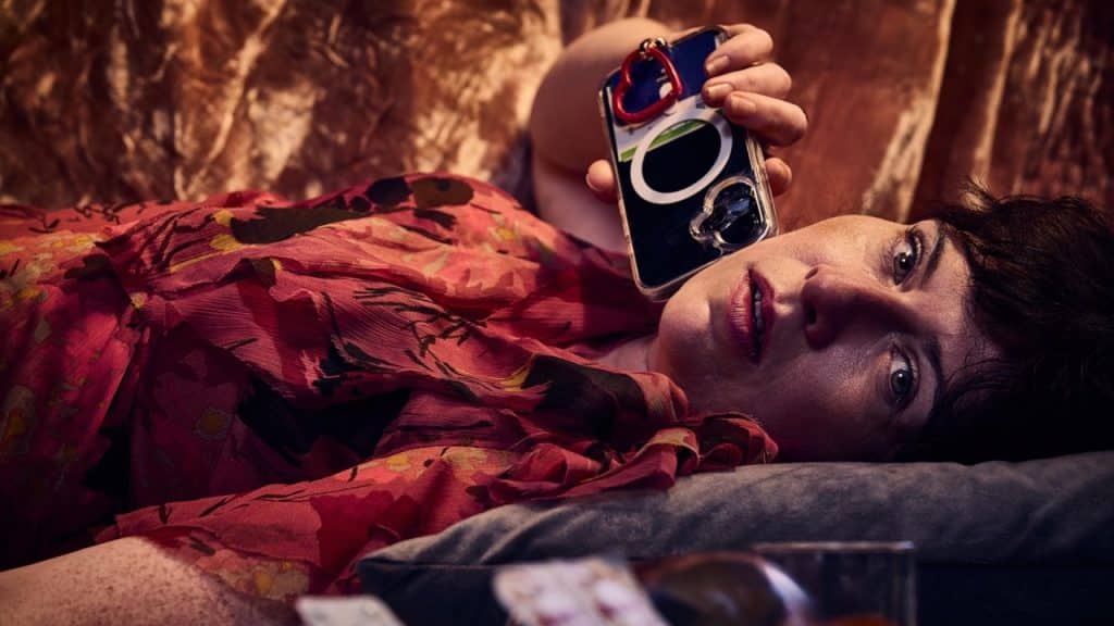 Actor Nicky Harley reclines against cushions in a richly coloured setting, holding a mobile phone close to her face. Dressed in a vivid floral blouse, she gazes directly toward the viewer.
