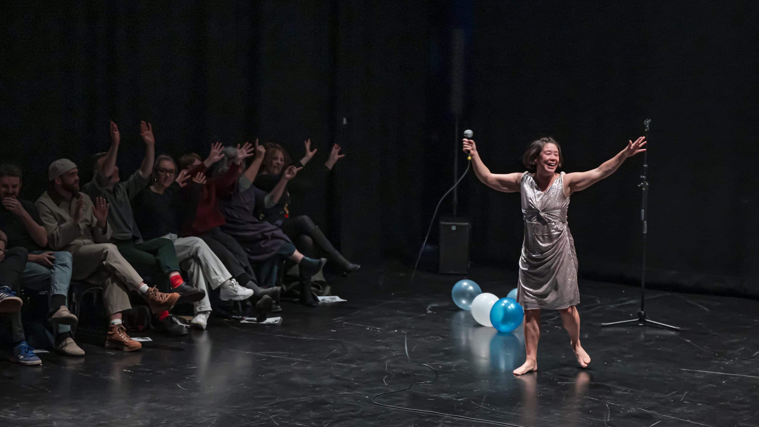 A picture of a live performance of The Rest of Our Lives from 2025. A barefoot performer in a silver dress stands centre stage, arms outstretched in a moment of joyful release, holding a microphone. To one side, a row of audience members sit close to the action. Balloons rest on the dark stage floor, suggesting a celebratory, intimate performance atmosphere.
