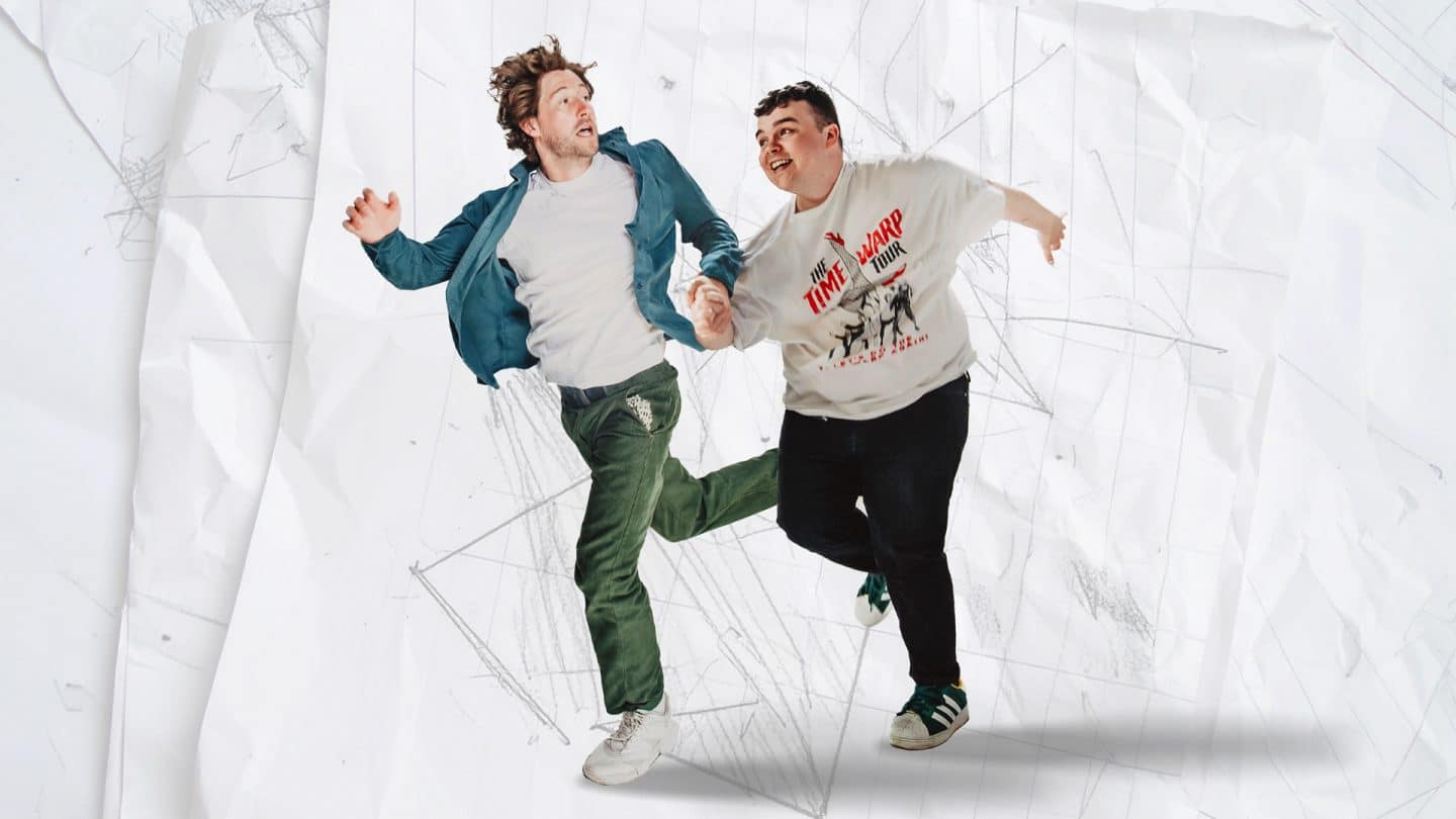 Two men are mid-jump, holding hands and smiling. One wears a white T-shirt with “The Time Warp Tour” printed on it and dark trousers; the other wears a white T-shirt, green trousers, and an open teal shirt. The background is a crumpled sheet of white paper with faint pencil sketches and grid lines.