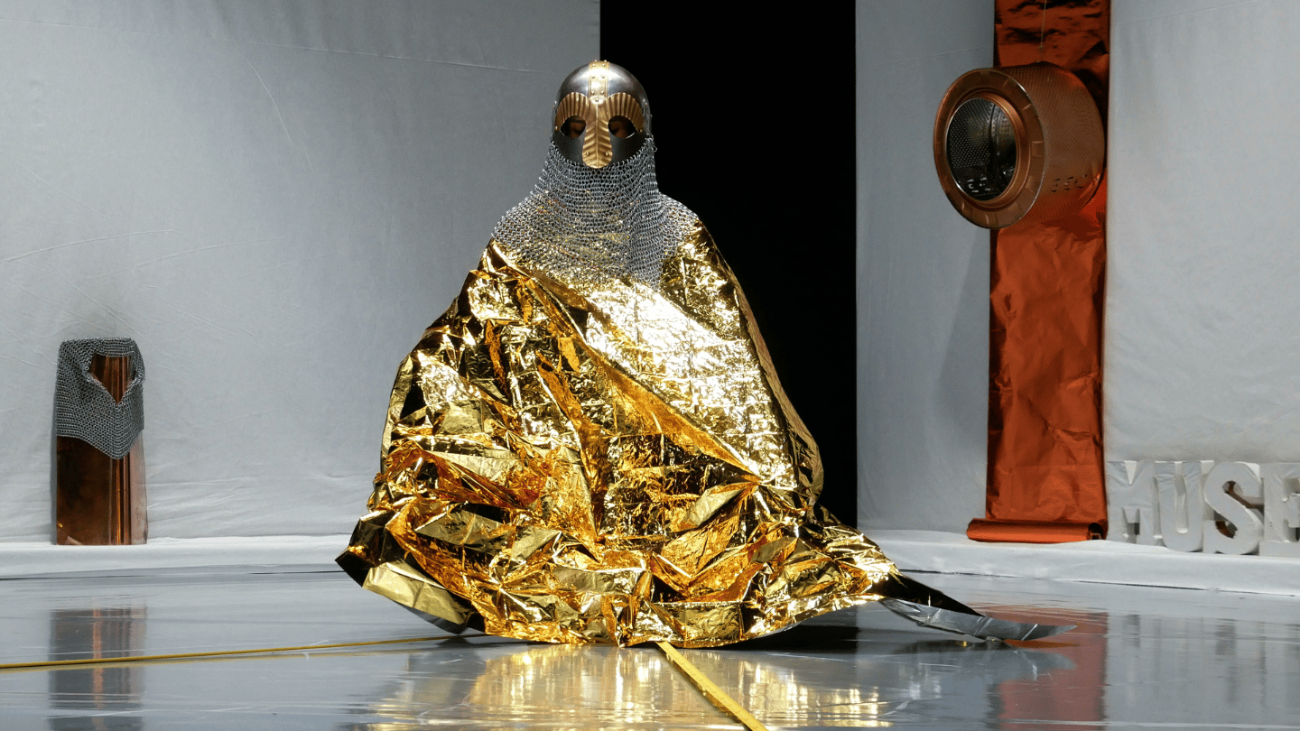 Untitled 14km. The setting is a museum gallery. In the centre, a human shaped figure appears like a sculpture, wearing a medieval style metal helmet and chainmail, over a gold foil cloak.