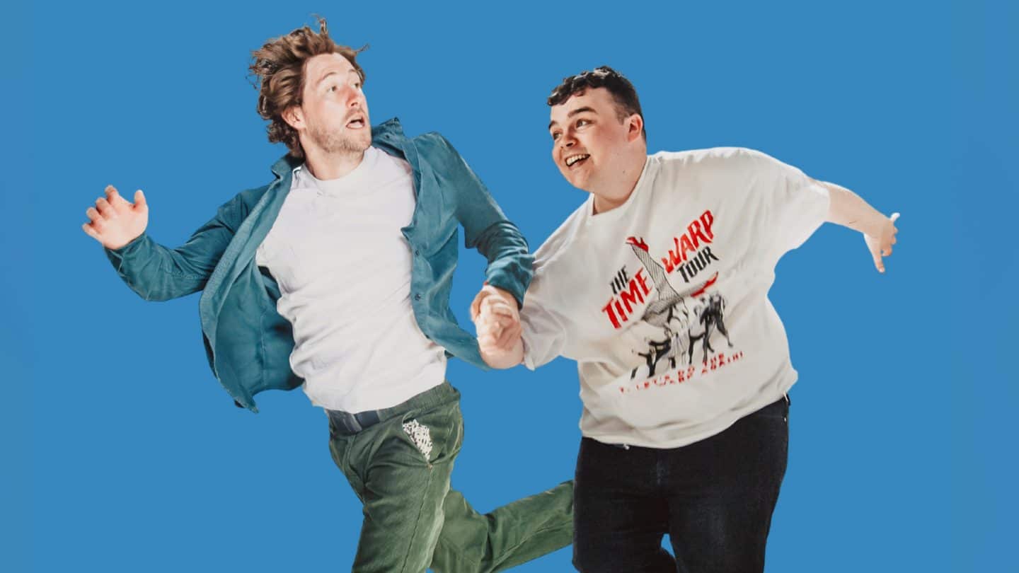Two men appear mid-jump against a bright blue background, holding hands.
