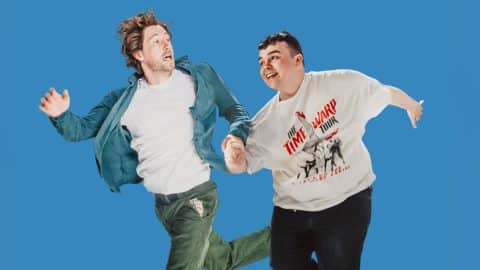 Two men appear mid-jump against a bright blue background, holding hands.
