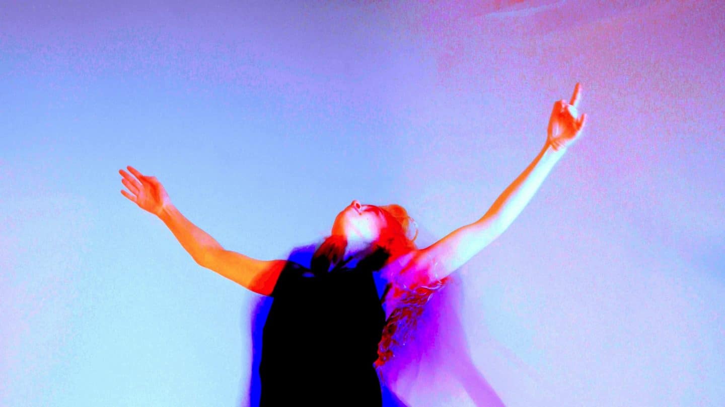 A woman in a black outfit stands against a gradient background of purple and pink hues, arms stretched wide and head tilted back in an expressive, open pose. The image is digitally altered with high-contrast and neon-like colours, giving it a surreal effect.